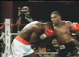 Sports gif. Mike Tyson is fighting in the ring with another boxer and he lands a huge uppercut, knocking the other boxer out. The text, "Boom!" flashes across the screen.