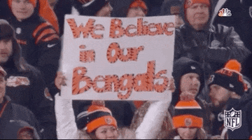 Sports gif. Amidst a sea of fans in black and orange Bengals winter gear, a woman with a Bengals beanie proudly holds up a sign that reads in tiger-striped letters, "We Believe in Our Bengals."