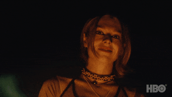 TV gif. Hunter Schafer as Jules in Euphoria. She stands behind a bonfire and the light from the flames illuminate across her face as she gives us a small, sad smile.