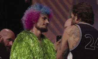Sports gif. UFC fighter Sean O'Malley is being interviewed by a person wearing a jacket made of neon green tinsel strands and a pink-purple clown wig. The interviewer asks, "You know what's gonna happen. You do huh?!" O'Malley responds, looking dead serious, with "We both know."