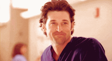 TV gif. Patrick Dempsey as Dr. Derek Shepherd on Grey’s Anatomy shakes his head and fights a smile on his face as he looks down at someone lovingly. 