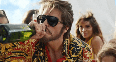 Music video gif. A man in a Taylor Swift music video swigs from a champagne bottle and sticks his tongue out while shaking his head. Girls in the back are dancing and partying with him.