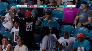 Video gif. Older man in an Atlanta Falcons jersey stands up and bows in an exaggerated way to the Miami Dolphins fans seated around him who are smiling awkwardly in response. 