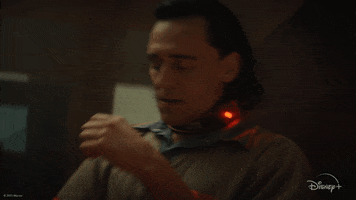 Disney gif. Tom Hiddleston as Loki on the show Loki sits in a prison jumpsuit. He leans back in his chair and rests his hand on his temple in frustration. 