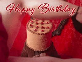 Video gif. We look through a woman's lifted leg and a man with a birthday cake on his head is slowly unbuttoning his shirt. Text, "Happy Birthday!"