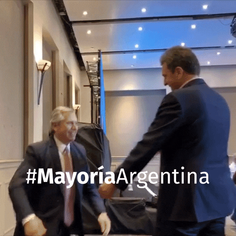 Political gif. Mayor of Argentina, Alberto Fernandez goes in for a big hug on stage with cameras flashing all around them.