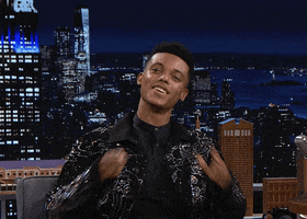Tonight Show gif. Jabari Banks, in a sparkly black jacket, brings his hands to his mouth and blows a grand kiss to the crowd, grinning.