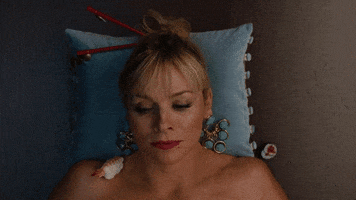 Video gif. A woman wearing chopsticks in her hair lies on a blue pillow, with sushi on her collarbone. She pops a piece of sushi in her mouth and chews.