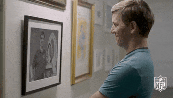 Sports gif. Eli Manning stands in a hallway smiling at a framed photo of him holding the NFL Pro Bowl Trophy. He looks at us with a cheeky smile, winks, and raises his eyebrows.