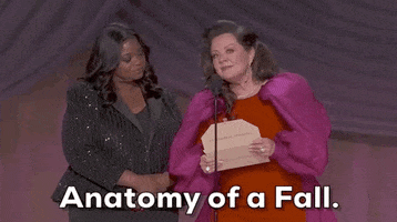 Oscars 2024 GIF. Melissa McCarthy and Octavia Spencer are reading the winner of Best Screenplay. McCarthy wears a red dress with big, puffy magenta sleeves, and Spencer wears a striped sequined black suit. McCarthy holds an open envelope and says, "Anatomy of a Fall" while Spencer's jaw drops next to her. 