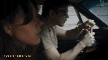 Movie gif. Swerving in a red Mustang, Aubrey Plaza as Sarah and Josh Hartnett as Danny in Operation Fortune: Ruse de Guerre frantically try to outrun a group of men shooting rifles from a black jeep.