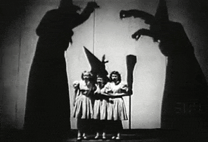 Video gif. Vintage black-and-white video of three huge creepy witch silhouettes as they dance around three little girls in white dresses.