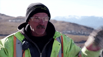 Reality TV gif. A man on Jade Fever wears a hazard jacket and thick gloves. He raises his hands and looks at his fingers as he tries to cross them. Text, "Fingers crossed."