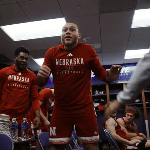 Sports gif. CJ Wilcher, a player on the Nebraska Cornhuskers basketball team, dances in the locker room with his teammates. He puts his hand on the back of his head and bounches his body with a big smile on his face.