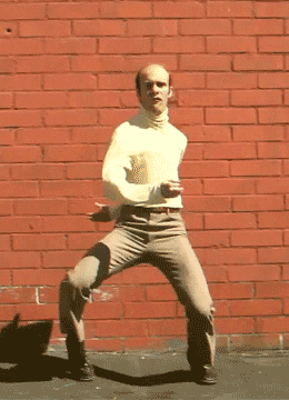 Video gif. Bald man wearing a turtleneck does a goofy dance in front of a red brick wall, flinging his arms awkwardly and stomping his feet as he holds a serious look in our direction.