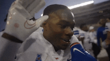 Sports gif. Closeup of Roderick Daniels from the SMU Mustangs football team smiling and waving at us as a teammate hangs off his shoulder on the sidelines.