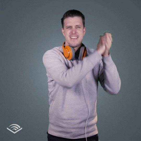 Video gif. A man in a light gray hoodie with orange headphones around his neck cheers us on with an old-timey clasped-hand victory gesture.