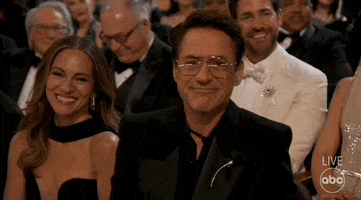 Oscars 2024 GIF. Robert Downey Junior, seated at the Oscars, looks at us and nods his head impishly, tapping his nose to confirm, Susan Downey beside him, shrugging and nodding in agreement.
