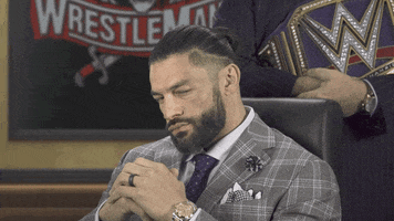Sports gif. Roman Reigns from WWE is wearing a suit and is sitting at his desk with his hands together. He stares us down and lifts his chin up, challenging us to approach.