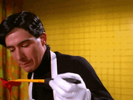 Music video gif. Ad-rock from the Beastie Boys in the music video Body Movin' wears an apron and comically large gloves. He looks straight into the camera with seductive eyes. He lifts a large, yellow fork dripping with a red sauce and licks it. Ad-rock continues to stare into the camera as he licks his lips.