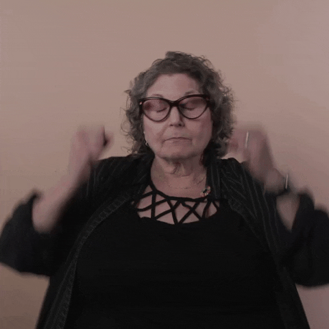 Reaction gif. A Disabled white woman with kinky curly gray hair and big wine-colored cat-eye glasses explodes her hands on each side of her head and puffs her cheeks out, gesturing "mind blown."