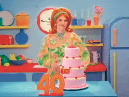 TV gif. A woman wearing a red 50s style wig and a pink, green, and yellow floral dress stands with her hands on her hips in front of a 40th birthday cake. She points her finger and shrugs her shoulders, giving a pleasant smirk. Text, "Happy birthday."