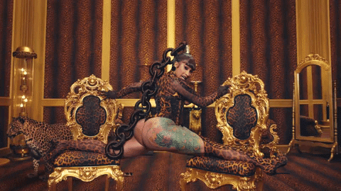 An animated gif from the music video WAP where Cardi B. is wearing a leopard print leotard, doing the splits between two ornate leopard print chairs, in a rich-looking room with leopard-print walls.