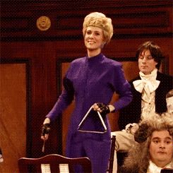 SNL gif. Kristen Wiig is wearing exercise clothes and holding a triangle, the percussion instrument. She smiles cheesily and kicks one leg into their air while staring at us.