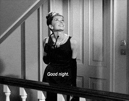 Movie gif. Audrey Hepburn as Holly Golightly in Breakfast at Tiffany's stands outside of an apartment, looking up and making a kissy face and then saying, "good night."