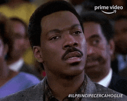 Movie gif. Eddie Murphy as Prince Akeem in Coming to America sits in a crowd of people. He looks straight ahead and looks away confused. He blinks quickly as if processing what he’s just heard. 