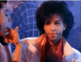 Music video gif. Wearing a white jacket with a popped collar, a big-haired Prince is about to take a sip from a mug, but is briefly taken aback by something he heard.