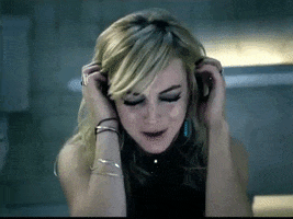 Music video gif. Lindsay Lohan in Confessions of a Broken Heart (Daughter to Father) covers her ears as she looks down in pain or sadness, singing or screaming. 