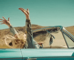 Music video gif. Kesha is in a convertible for her music video for I Need a Woman to Love. Her hands are both in the air and her hair is flying behind her as she cruises through the desert.