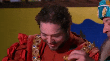 Tonight Show gif. Post Malone wears old-fashioned, royalty clothes as he holds a piece of food in his hand. He clinks his piece of food with Jimmy Fallon’s piece of food like they’re clinking glasses. They then both take bites out of the food.