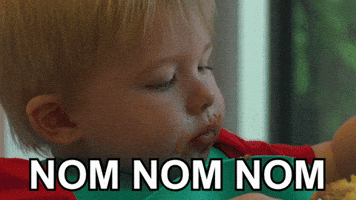 Video gif. Baby with a bib on is eating and begins to feed himself with his left fist but sees his right fist holding a fork coming to his mouth first, so he switches at the last minute and takes a big bite. Text, "Nom nom nom."