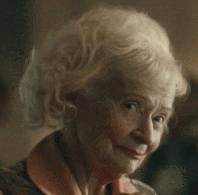 Video gif. An elderly lady looks coyly at us while slowly looking up and down, and she has a sly smile on her face.