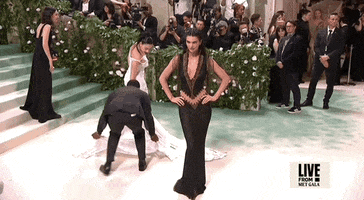 Met Gala 2024 gif. Kendall Jenner wearing a plunging black and bronze vintage Givenchy 1999 Haute Couture gown and posing for the cameras, drops her hands from her hip and snaps into it.