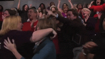 Video gif. A grainy clip shows a crowd going wild with applause as two women in the front jump up and down while hugging each other.