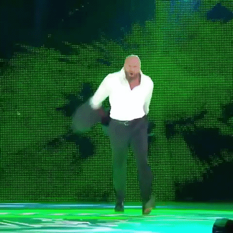 Video gif. Man takes off his suit jacket and loosens his collared shirt, running across a stage to meet a waiting opponent in a boxing rink.