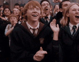 Movie gif. Rupert Grint as Ron Weasley in Harry Potter claps and grins alongside a crowd of cheering, applauding Hogwarts students.