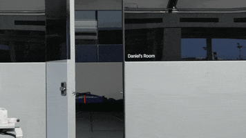 Sports gif. Daniel Ricciardo in his racing suit appears in a doorway, smiles and waves at us, then leaves to the right. Text printed next to the doorway marks the area as “Daniel’s Room.”