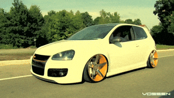Video gif. A white Volkswagen GTI with bright yellow rims drives down a road lined with lush, green trees. The driver's side window is rolled down.