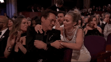 Oscars 2024 GIF. Emily Blunt leans over the row to put her arms around Robert Downey Junior and share a laugh with him.
