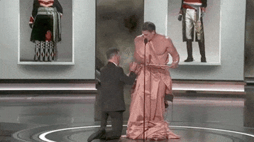 Oscars 2024 GIF. Jimmy Kimmel is kneeling down in front of John Cena as he helps Cena adjust the robe made out of a curtain that Cena has put on. Finally clothed, Cena opens the envelope and says, "And the Oscar goes to..."