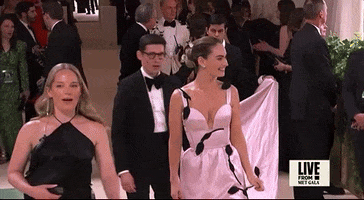 Met Gala 2024 gif. Lily James waves and smiles as she makes her entrance. She's wearing a pale pink Erdem gown with a fitted bodice, long train, thin straps, and scalloped plunging neckline. A black vine with leaves detail weaves across the skirt and over her shoulders. 
