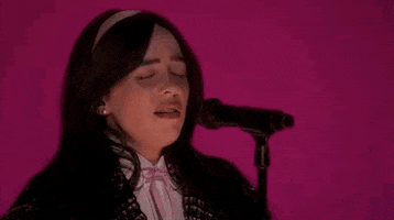 Oscars 2024 GIF. Billie Eilish performing "What Was I Made For" on stage at the Oscars. Close up zoom on Eilish as she croons into the microphone, her face contorting with passion, effort, and feeling. The lighting behind her is glowing with Barbie pink. 