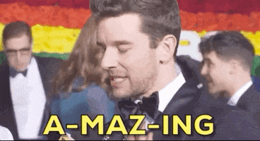 Video gif. In front of a rainbow flag background, a stubbly man in a tuxedo faces left of frame. He bounces as he speaks, his eyes widen, and his lips emphasize the words: Text, "A-maz-ing."