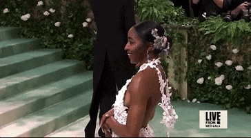 Met Gala 2024 gif. Closeup of Ayo Dibiri swaying back and forth in a white halter backless Loewe gown covered in a textured lace pattern. The halter neckline arcs over the top of her shoulders, hanging down in a loop at her back.