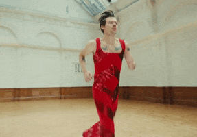 Music video gif. Harry Styles in his video for As It Was wears a red sequined jumpsuit as he jogs wearily in a cavernous ballroom.