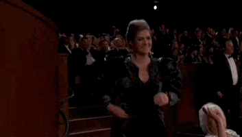 Oscars 2024 GIF. Holly Waddington briskly walks down the aisle to accept her Oscar, diverting briefly to give us two thumbs up.